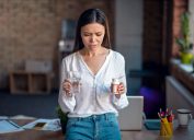 A young woman who's not feeling well leans against her desk with a glass of water in one hand and a pill bottle in the other