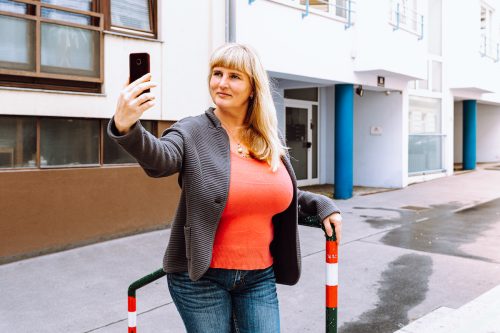 A happy woman wearing jeans, an orange t-shirt, and a gray blazer, takes a selfie on a city street