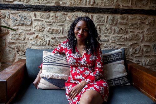 A mature woman wearing a red dress with a white floral print sits on a couch in front of a stone wall