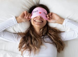 Top view of a happy woman wearing a funny pink sleeping mask enjoying morning lying in comfortable bed