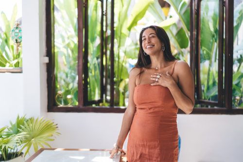 A young woman wearing an orange dress is smiling and touching her chest while in a tropical location