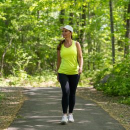 A middle-aged woman wearing black leggings and a bright yellow tank is taking a walk along a tree-lined path