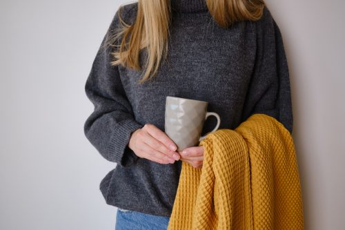 Close up of a woman dressed in a dark gray sweater holding a mustard-yellow blanket and a mug