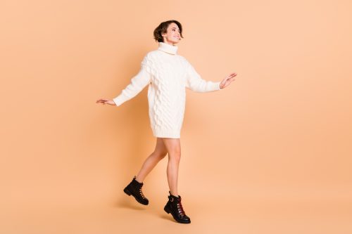 Young woman wearing white sweater dress with black boots against a peach background