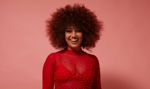Woman with an afro wearing a sheer red top against a pink background.