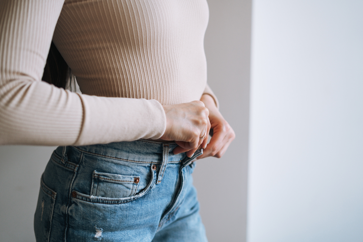 Close up of a woman's midsection as she buttons her jeans with a beige top.
