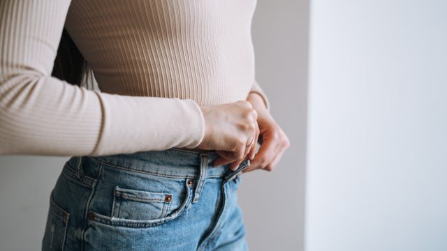Close up of a woman's midsection as she buttons her jeans with a beige top.