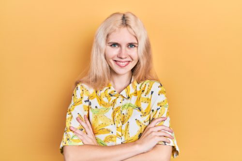 A young blonde woman wearing a banana-print shirt poses with her arms crossed in front of a yellow background