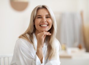 Happy attractive blonde woman in white silky bathrobe posing at white bathroom, smiling at camera, closeup portrait, panorama with copy space. Face and skin care at home concept