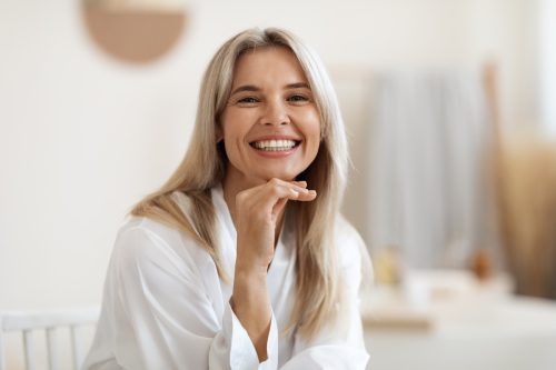 Happy attractive blonde woman in white silky bathrobe posing at white bathroom, smiling at camera, closeup portrait, panorama with copy space. Face and skin care at home concept