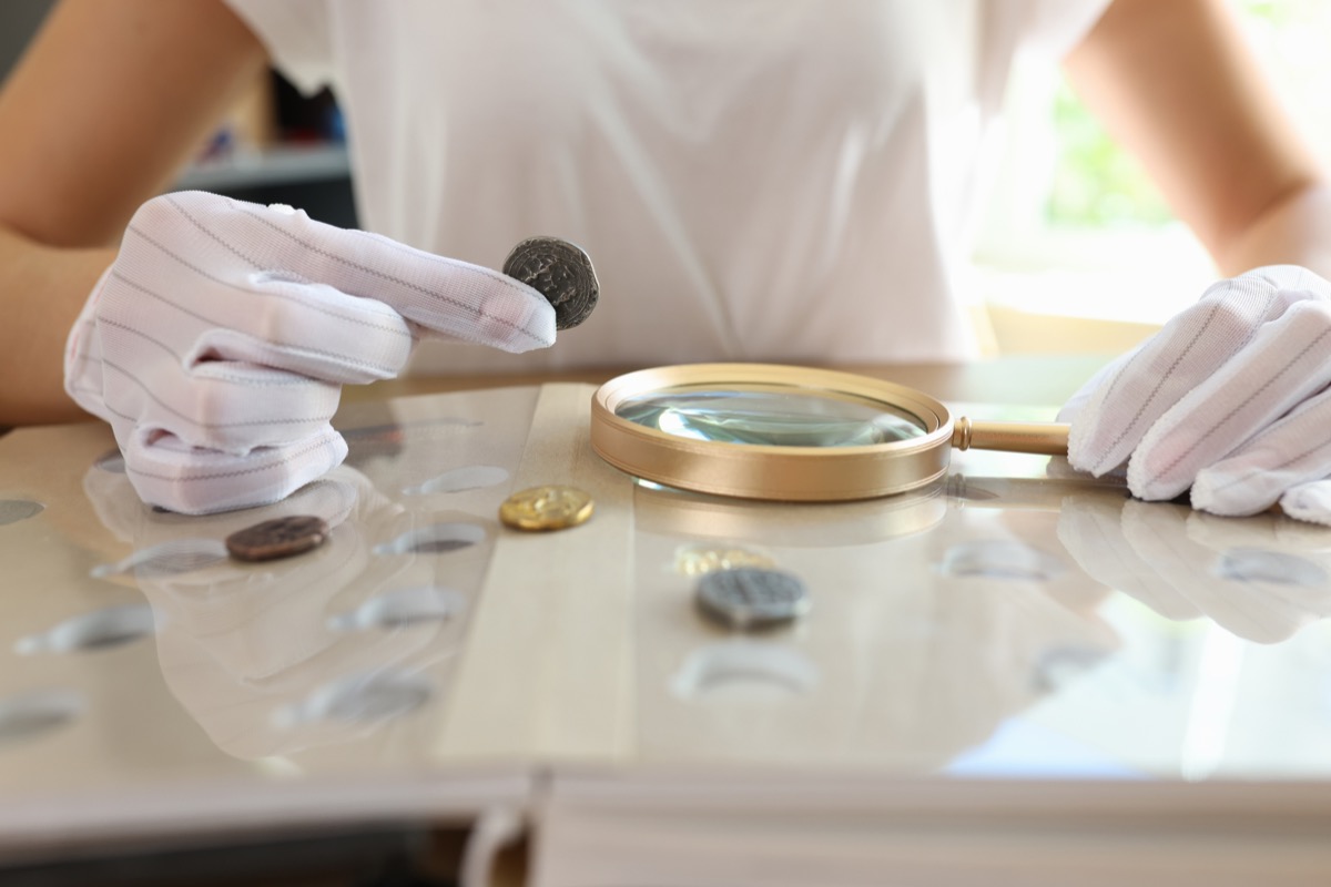 Female numismatist examines collection of coins. Woman looks at coins through magnifying glass. Numismatic collection review.