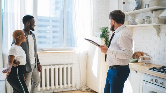 Side view of professional bearded chubby man in formal outfit with clipboard inspecting kitchen together with inspired ethnic couple while standing at modern sunny apartment