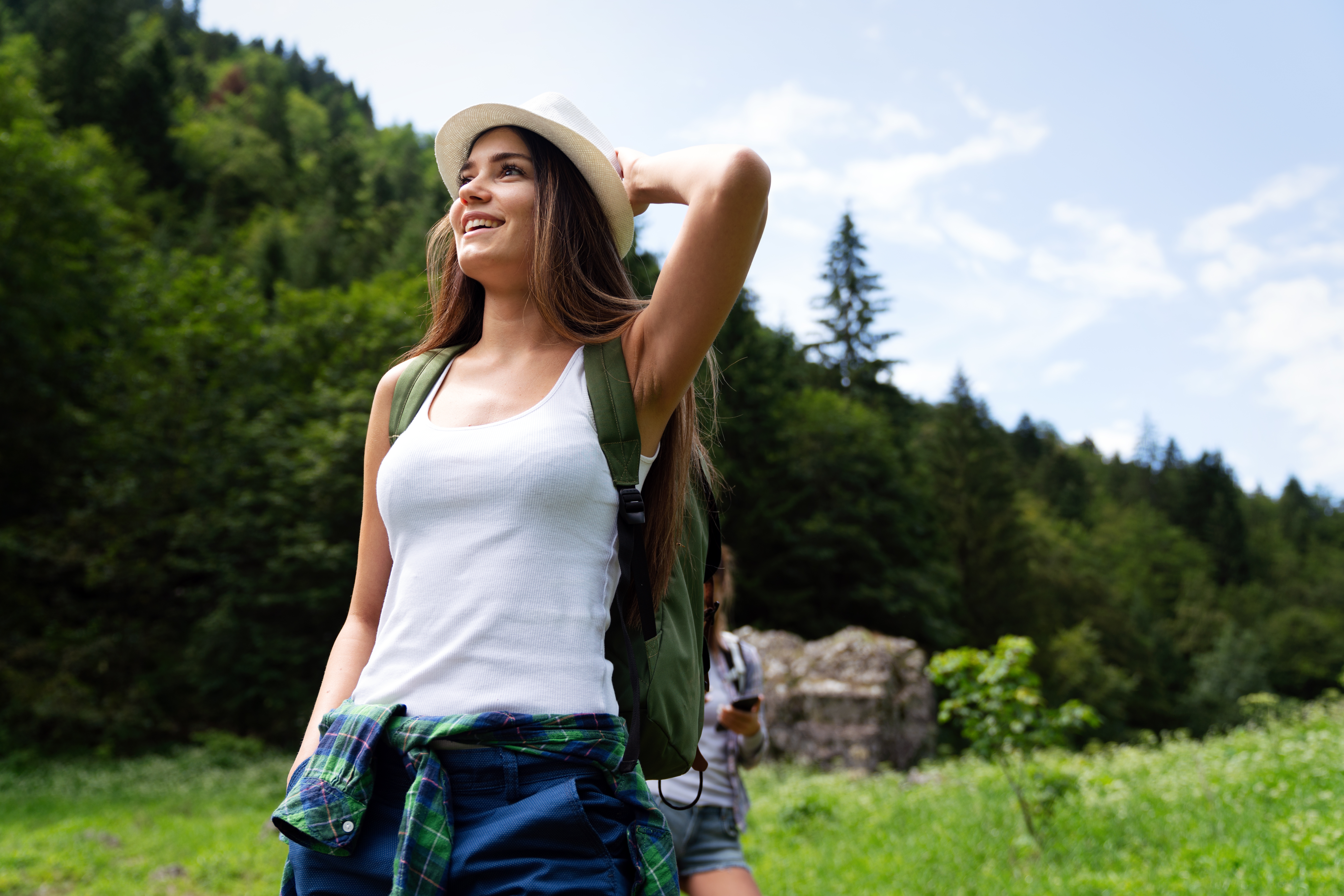 Happy young woman wearing a white tank top, hat, and green backpack standing in a forest while on a hike