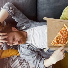 Above view portrait of bearded man lying on couch and eating pizza while watching TV at home, copy space