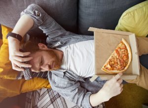 Above view portrait of bearded man lying on couch and eating pizza while watching TV at home, copy space