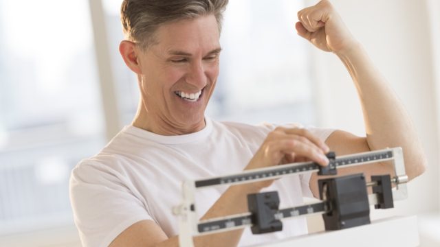 Excited mature man clenching fist while using balance weight scale at gym