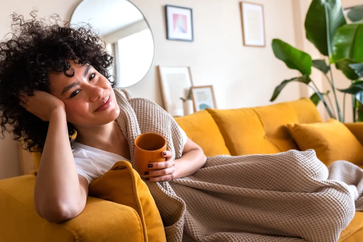 Happy woman relaxing at home lying on couch holding cup of warm coffee looking at camera.