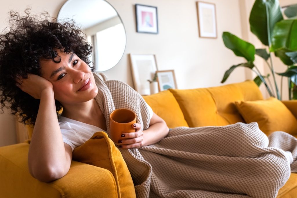 Woman smiling at camera, laying on couch with blanket and mug