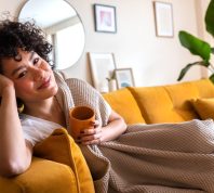 Happy African American woman relaxing at home lying on couch holding cup of warm coffee looking at camera. Copy space.