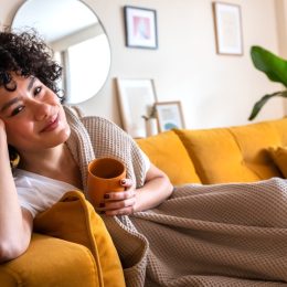 Happy African American woman relaxing at home lying on couch holding cup of warm coffee looking at camera. Copy space.
