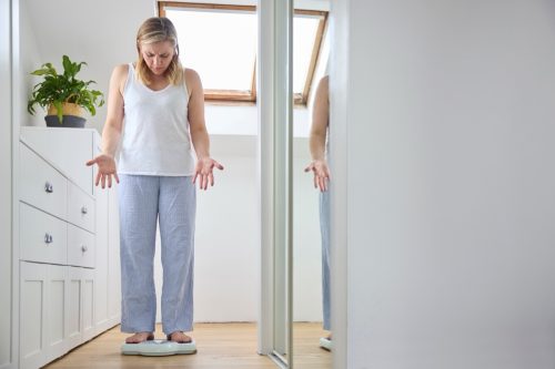 Menopausal Mature Woman Concerned With Weight Gain Standing On Scales In Bedroom At Home