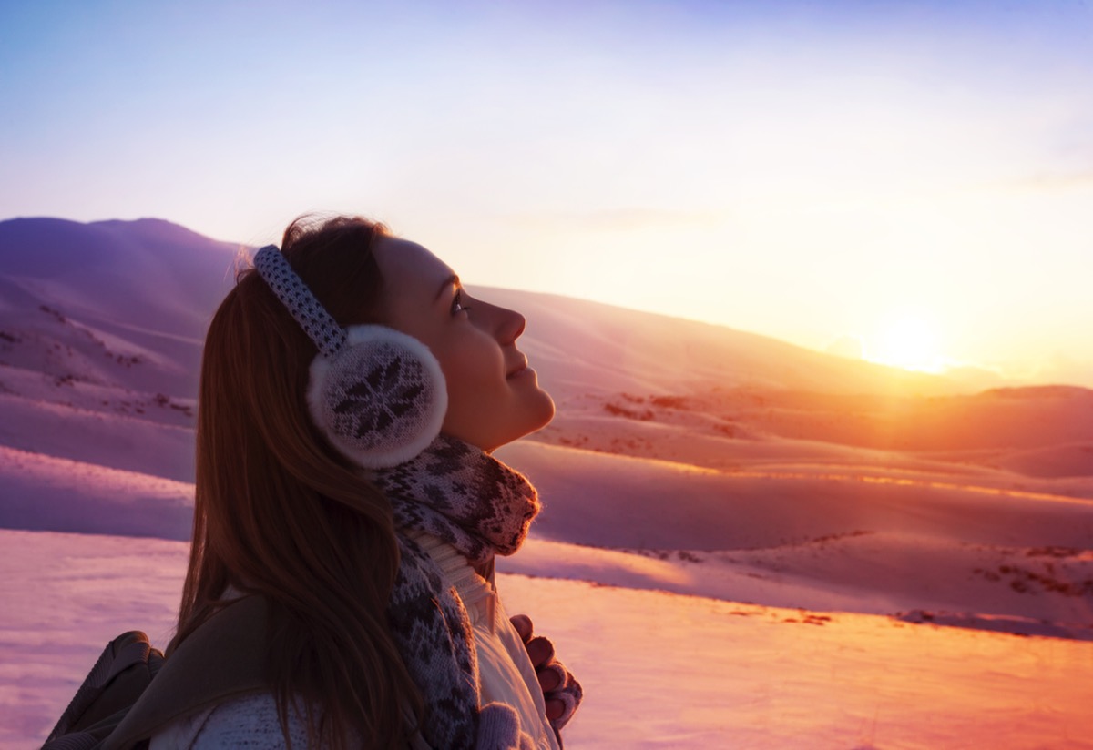 Image of pretty woman walking in snowy mountains, side view of cute girl looking up, closeup portrait of female wearing warm winter earmuff, red sunset, wintertime sports, trekking and hiking concept
