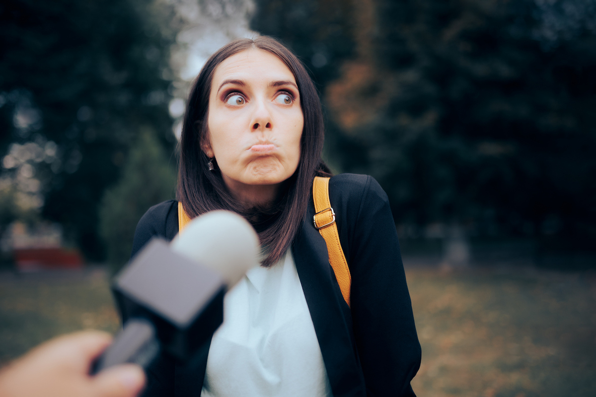 A young woman standing outside and looking awkward while a microphone is pointed towards her.