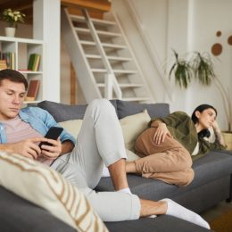 bored young couple watching TV on sofa at home, focus on man using smartphone in foreground