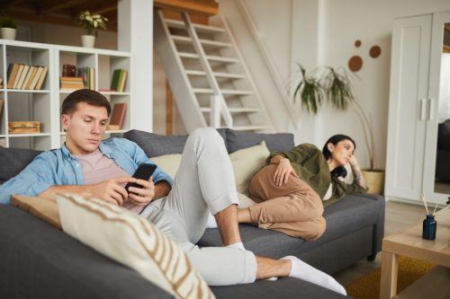 bored young couple watching TV on sofa at home, focus on man using smartphone in foreground