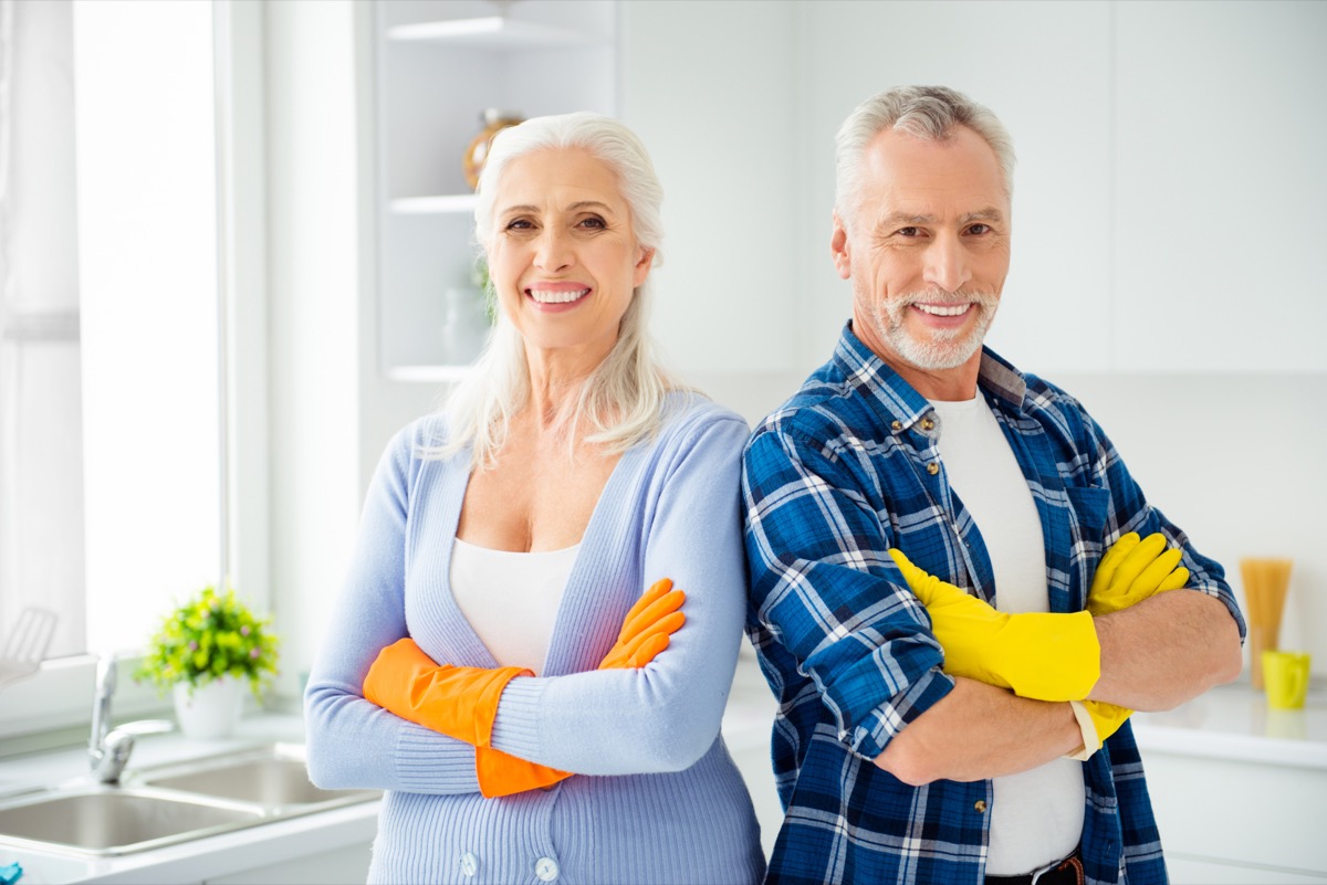 Lovely attractive cleanly neat cheerful stylish couple of senior in colorful gloves standing with crossed arms after cleaning, looking at camera, in the kitchen flat apartment