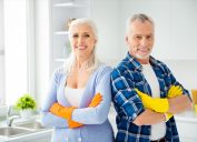 Lovely attractive cleanly neat cheerful stylish couple of senior in colorful gloves standing with crossed arms after cleaning, looking at camera, in the kitchen flat apartment