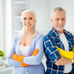 Lovely attractive cleanly neat cheerful stylish couple of senior in colorful gloves standing with crossed arms after cleaning, looking at camera, in the kitchen flat apartment