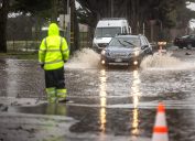 A traffic guard in hi-vis gear guides a car away from a flooded area in a street