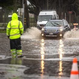 A traffic guard in hi-vis gear guides a car away from a flooded area in a street