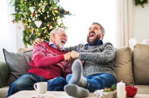 A senior father and adult son sitting on a sofa at home at Christmas time, laughing.