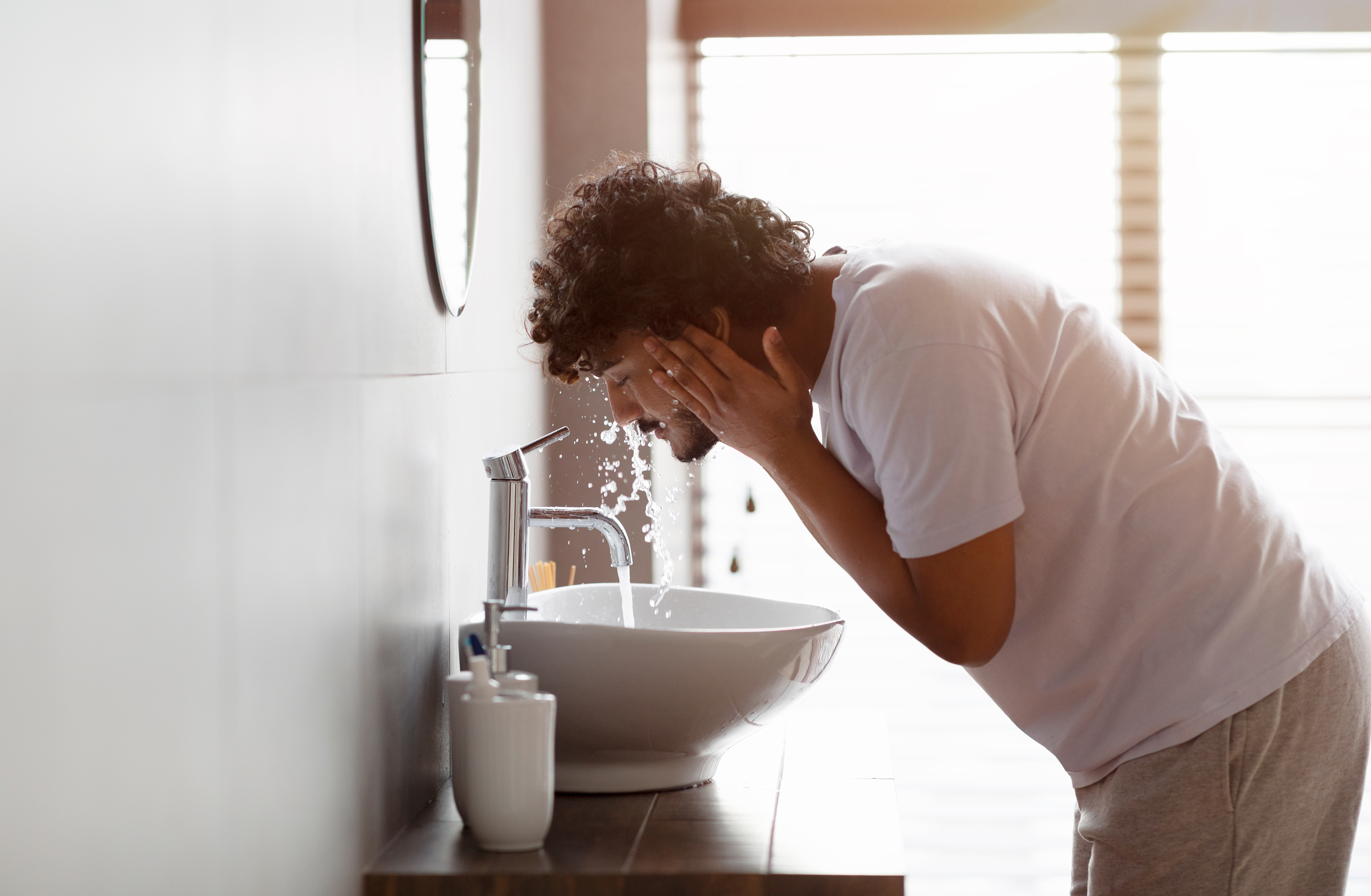 A man washes his face in a hotel room sink at dawn.