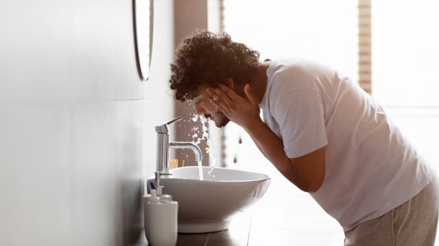 A man washes his face in a hotel room sink at dawn.