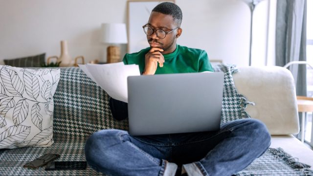 Young casually clothed concentrated man going over paperwork while working from living room, he is sitting on the sofa with laptop in his lap