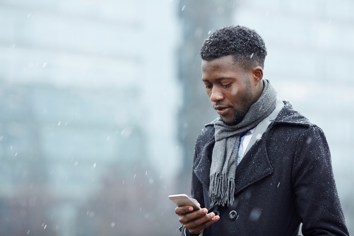 Portrait of handsome man using smartphone phone in snowy autumn city street, typing text message and looking at screen