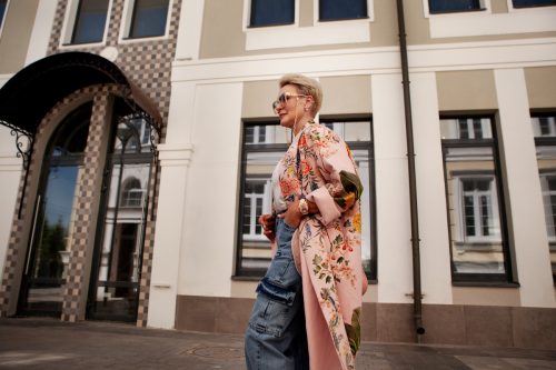 Stylish mature woman walks near city building, wearing cargo denim pants, pink floral trench coat