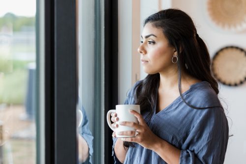 The mid adult woman takes a break with a hot drink in a mug. She is standing at the sliding glass doors and thinking.