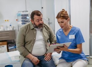 Female doctor consulting with the overweight patient, discussing test result in doctor office. Obesity affecting middle-aged men's health. Concept of health risks of overwight and obesity.
