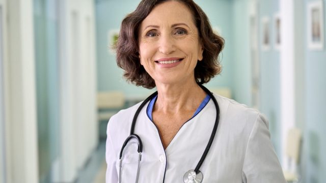 Portrait of smiling older senior female professional doctor physician pediatrician wearing white robe with stethoscope around neck standing in modern private clinic hospital, looking at camera.