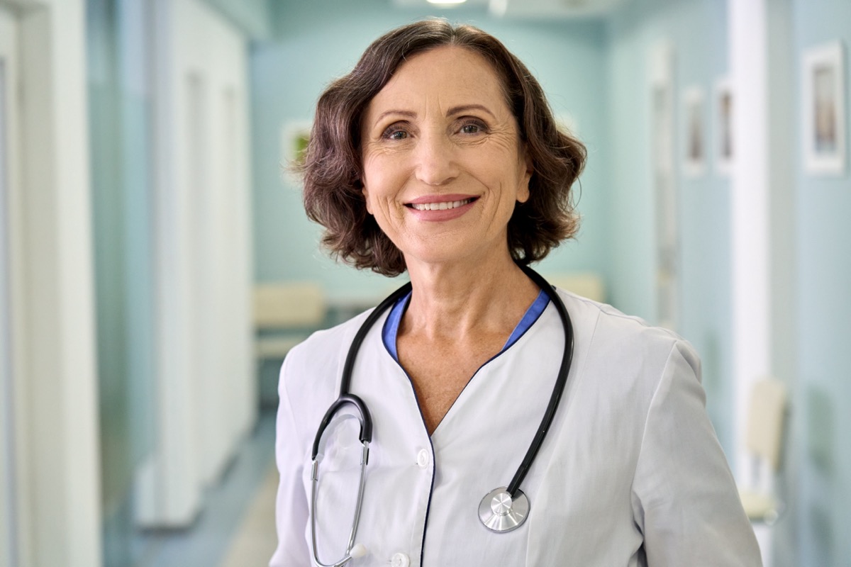 Portrait of smiling female doctor wearing white coat with stethoscope around neck standing in modern private clinic hospital, looking at camera.