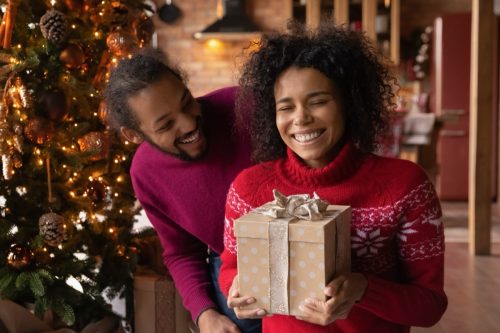 man and woman laughing in front of the Christmas tree while unwrapping presents