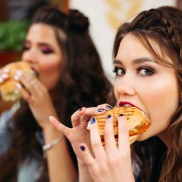 Close-up of beautiful teen girls with bright professional make up taking a bite of delicious crunchy burger and looking at camera.