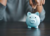 Blurred image of a person wearing a blue sweater putting a coin into a blue piggy bank.