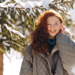 Smiling woman in winter clothes looks at the camera and puts her hair behind her ear. A young red-haired lady poses against the backdrop of snow-covered trees during a daytime walk in the park.