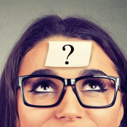 woman in glasses with a question mark post-it note on her forehead looking up over a gray background
