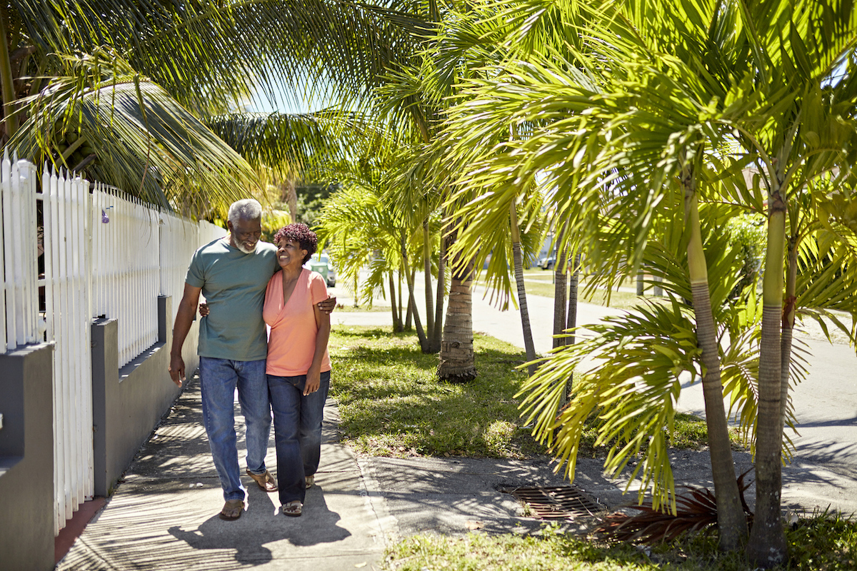 A senior couple hugging and walking down a sidewalk lined with palm trees on a sunny day.